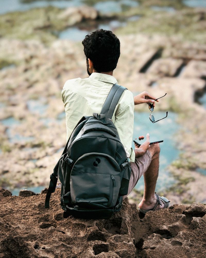 A man with a backpack sits on a rocky cliff overlooking a tide pool.