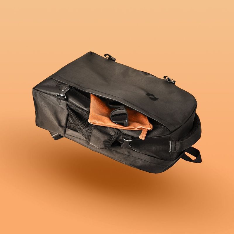 Black backpack floating against orange background with sunglasses and leather pouch inside open compartment.