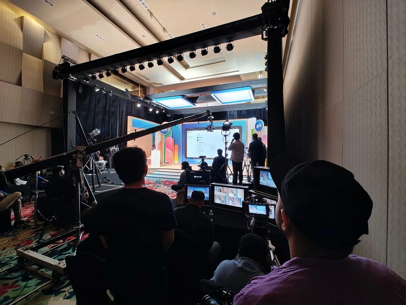 Production crew filming a stage event with cameras, monitors, and a large lighting truss.